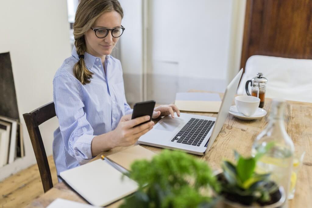 Woman Using Cell Phone and Laptop on Wooden Desk at Home.