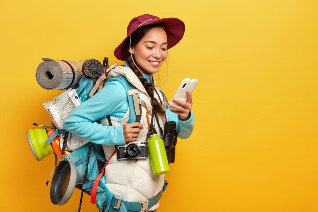 Positive pretty Asian tourist uses modern gadget for navigating, dressed in hat and jumper with jacket.
