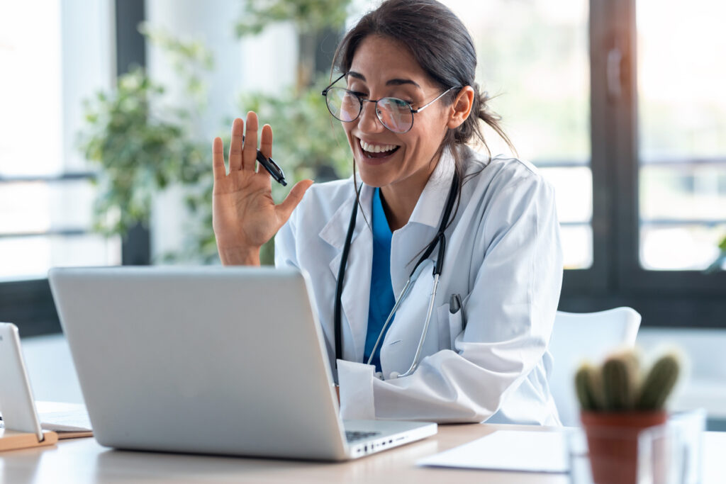 Virtual Medical Assistant Female Doctor waving and talking with patients through a video call with a laptop in the consultation.