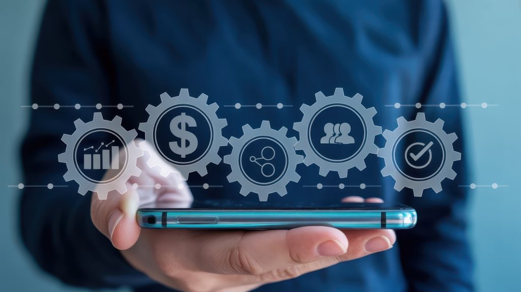Person holding a slim modern smartphone horizontally, projecting a holographic or augmented reality interface featuring five interlocked gears containing various business and process icons like a bar chart, a dollar sign, a share symbol, a group of people, and a checkmark, suggesting digital transformation, operational efficiency, or mobile business solutions.