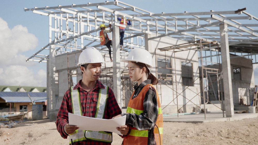 Construction engineers in hard hats and high-visibility safety vests review blueprints together on a modern residential home building site, with steel framework and workers assembling the structure in the background, illustrating the transition from design plans to advanced home construction using emerging technologies like modular building and digital planning.