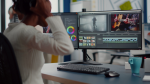 Female video editor working from home at a modern dual-monitor workstation, wearing headphones and focused on video editing software. The monitors display a timeline with video clips, color grading tools, preview of a dancing couple in motion, and a close-up of someone playing acoustic guitar. The desk setup includes a mechanical keyboard, mouse, blue coffee mug, and organized workspace, representing content creation, post-production, filmmaking, YouTube editing, and multimedia projects as a work-from-home creative professional.