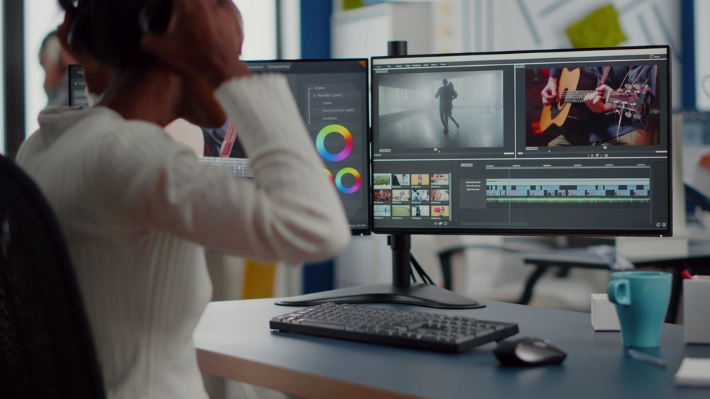 Female video editor working from home at a modern dual-monitor workstation, wearing headphones and focused on video editing software. The monitors display a timeline with video clips, color grading tools, preview of a dancing couple in motion, and a close-up of someone playing acoustic guitar. The desk setup includes a mechanical keyboard, mouse, blue coffee mug, and organized workspace, representing content creation, post-production, filmmaking, YouTube editing, and multimedia projects as a work-from-home creative professional.