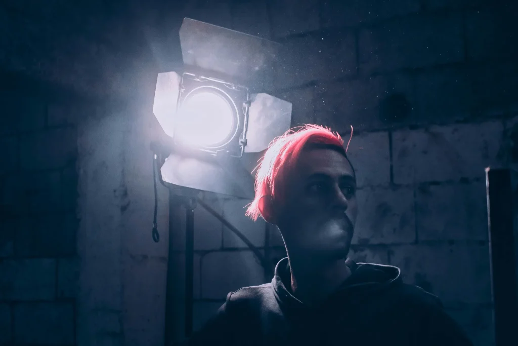 Dramatic presentation lighting setup featuring a person with neon red hair standing in a dark industrial environment beside a powerful studio spotlight. The cinematic blue-toned scene highlights how professional lighting influences audience focus, emotional engagement, visual storytelling, and presentation atmosphere during live events, conferences, and brand presentations.