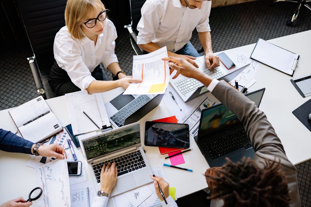 Employees collaborating in a technology-enabled workplace, analyzing reports, financial charts, and operational data using laptops and mobile devices. This modern office meeting illustrates data-driven decision making, workplace management, digital transformation, business intelligence, employee collaboration, hybrid work technology, and smart workplace solutions that improve productivity and organizational performance.